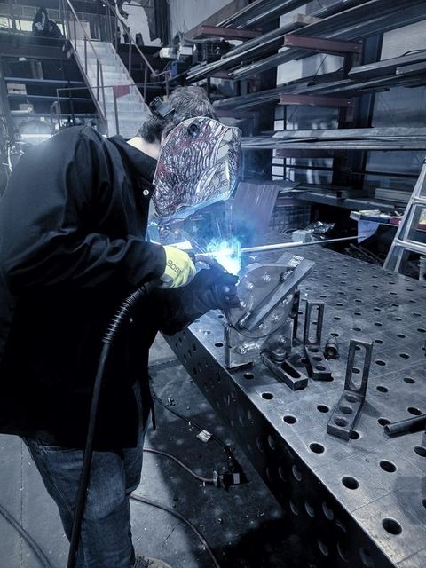 A welder in a painted hood welds a part on the fixture table
