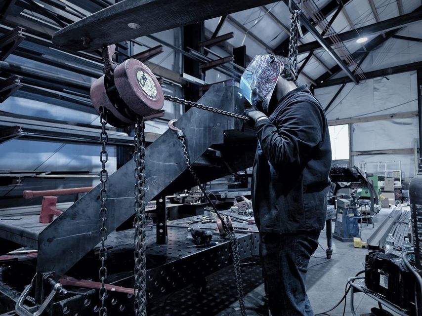 A welder in a hood works on a stair stringer hanging from a chain hoist