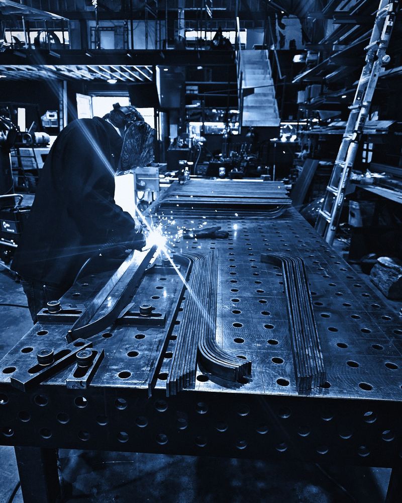 A welder strikes an arc over parts clamped to a long fixture table